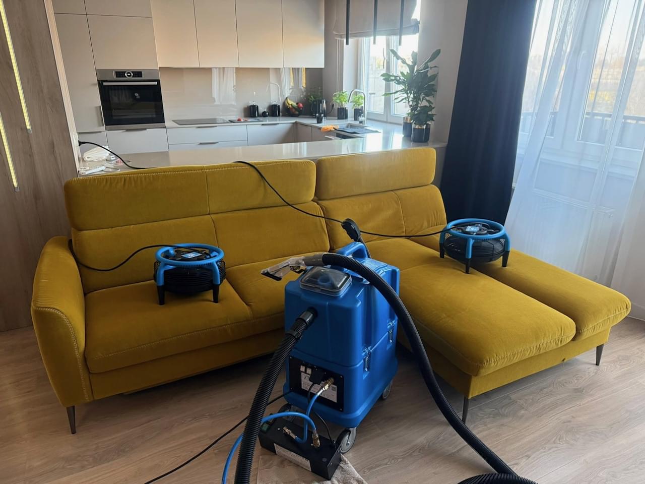 Proces profesjonalnego prania i suszenia żółtej kanapy narożnej za pomocą odkurzacza piorącego i dwóch osuszaczy.